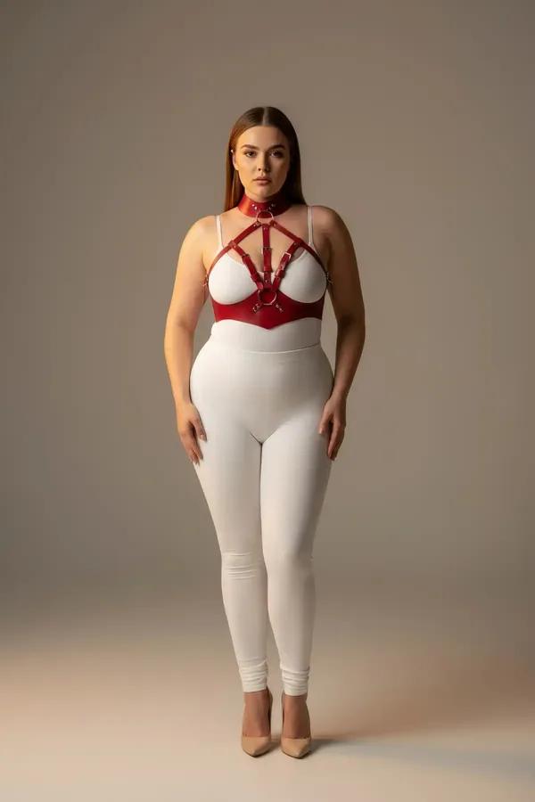 Red faux leather strappy chest harness with choker collar and gold O-rings worn over white bodysuit