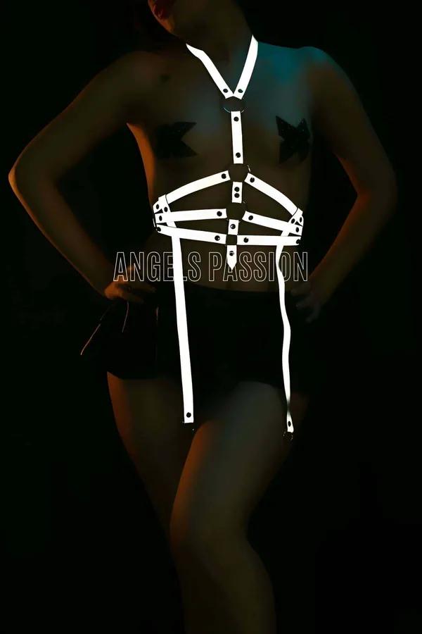 Reflective white-silver O-ring body harness with chest cage straps and dangling leg straps glowing under dark studio lighting