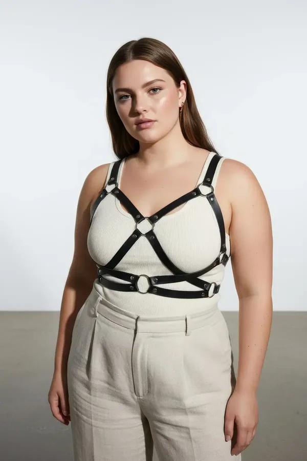 black faux leather criss-cross body harness with silver O-rings worn over cream ribbed tank top plus size model